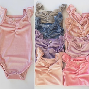May include: A set of seven velvet baby bodysuits in various shades of pink, purple, blue, and beige. Each bodysuit has ruffled straps and a gathered neckline.
