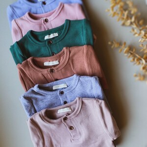 Puede incluir: Una pila de seis camisas de manga larga en varios colores, incluyendo azul claro, rosa, verde, marrón, morado y rosa oscuro. Las camisas tienen un solo botón en el cuello y una textura acanalada.