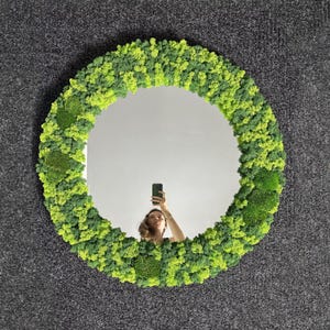 May include: A round mirror with a vibrant, textured frame made of green and yellow moss. The mirror reflects a person holding a phone. The moss frame adds a natural, organic element to the decor.