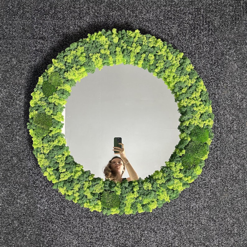 May include: A round mirror with a vibrant, textured frame made of green and yellow moss. The mirror reflects a person holding a phone. The moss frame adds a natural, organic element to the decor.
