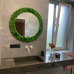 May include: A round mirror with a vibrant green moss frame hangs above a dark gray sink. The bathroom features gray tile walls, a window, and a countertop with a soap dispenser and a candle.