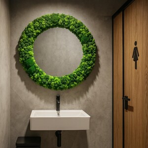 May include: A modern bathroom with a round mirror framed by green moss. A white rectangular sink with a black faucet is below the mirror. A black trash can sits to the left of the sink, and a wooden door with a female restroom symbol is on the right.