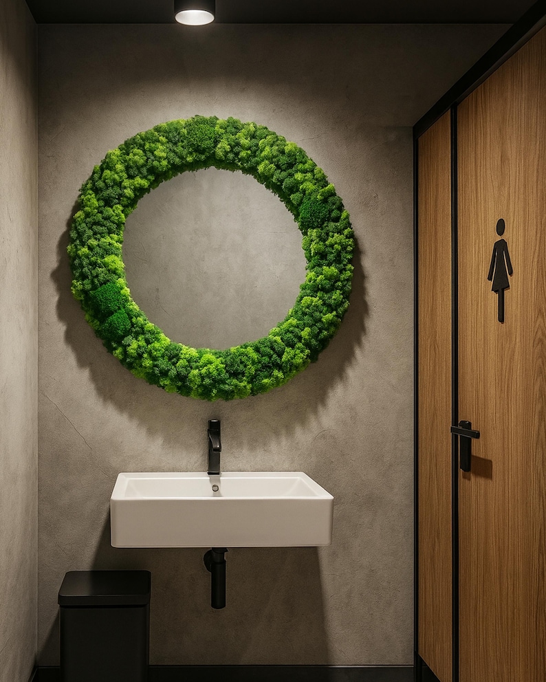 May include: A modern bathroom with a round mirror framed by green moss. A white rectangular sink with a black faucet is below the mirror. A black trash can sits to the left of the sink, and a wooden door with a female restroom symbol is on the right.