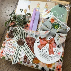 May include: A gift set featuring a floral-patterned package with a highland cow illustration, a green tag labelled Girl, two lavender pens, a striped ribbon with a brown button, a fabric bow, and stickers with text including With God all things are possible.