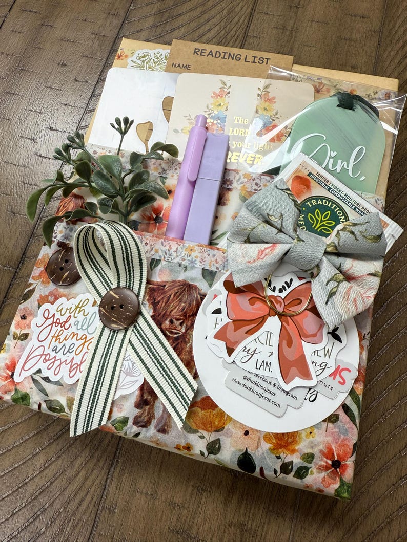 May include: A gift set featuring a floral-patterned package with a highland cow illustration, a green tag labelled Girl, two lavender pens, a striped ribbon with a brown button, a fabric bow, and stickers with text including With God all things are possible.