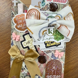 May include: A rectangular package wrapped in floral paper featuring a large fabric bow with a rainbow pattern. The package is decorated with various stickers including text like WWJD, Do Justice, and a candle illustration, plus small bows and buttons.