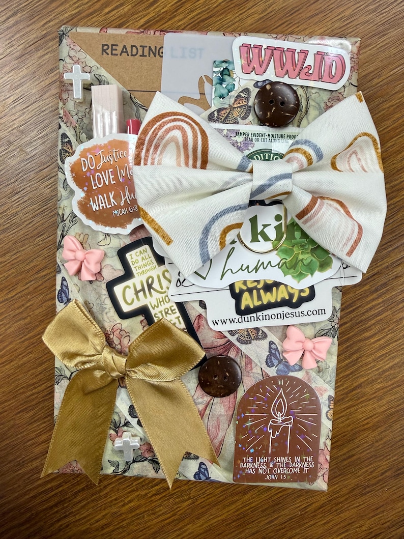 May include: A rectangular package wrapped in floral paper featuring a large fabric bow with a rainbow pattern. The package is decorated with various stickers including text like WWJD, Do Justice, and a candle illustration, plus small bows and buttons.
