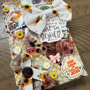 May include: A rectangular book cover wrapped in fabric with a sunflower and daisy pattern, decorated with a large sunflower bow, various stickers including a cow, a pig, and text reading "Keep Calm and Call on Jesus" and "Trust in God".