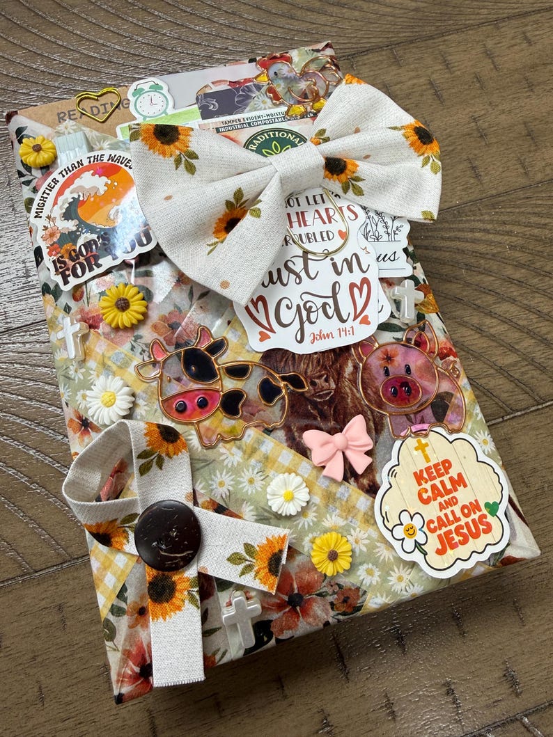 May include: A rectangular book cover wrapped in fabric with a sunflower and daisy pattern, decorated with a large sunflower bow, various stickers including a cow, a pig, and text reading "Keep Calm and Call on Jesus" and "Trust in God".