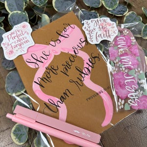 May include: A brown card featuring a pink ribbon illustration and the text She is far more precious than rubies Proverbs 31:10, accompanied by two stickers with floral designs and text, a floral bookmark, and a light pink pen with a moustache logo.