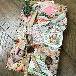 May include: A gift package wrapped in floral paper featuring a fabric bow, two brown buttons, and a tan ribbon. It includes a tea bag, a small artificial plant, a pink pen, and stickers with text including Let everything that has breath praise the Lord.