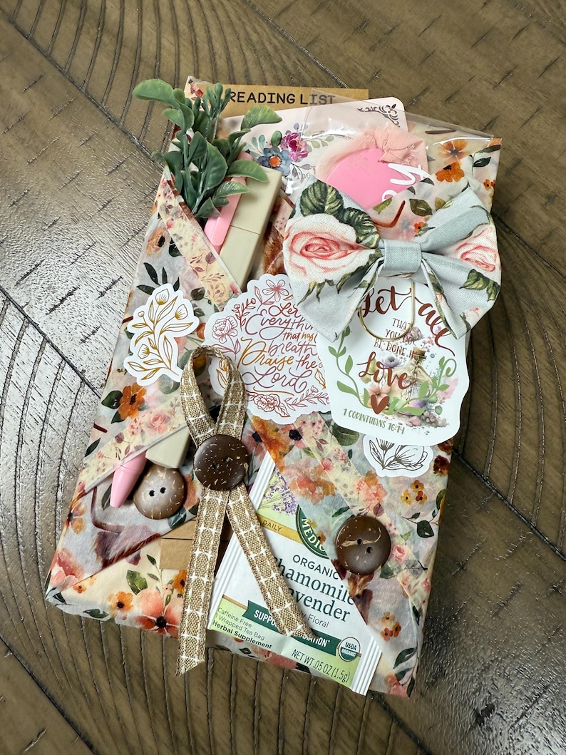 May include: A gift package wrapped in floral paper featuring a fabric bow, two brown buttons, and a tan ribbon. It includes a tea bag, a small artificial plant, a pink pen, and stickers with text including Let everything that has breath praise the Lord.