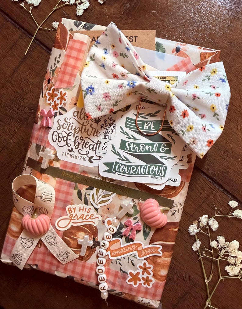 May include: A rectangular package wrapped in pumpkin-patterned paper, adorned with a floral fabric bow, a beaded string spelling REDEEMED, and various stickers featuring text like "all scripture is God-breathed" and "BE STRONG & COURAGEOUS."