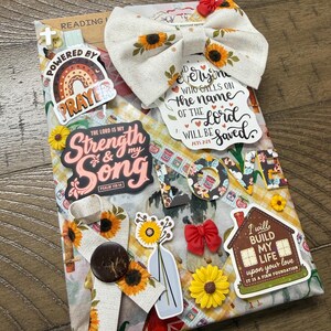 May include: A rectangular devotional book decorated with sunflower-patterned fabric bows, religious stickers, and cutouts. Text includes "Powered by Prayer," "The Lord is my Strength & my Song," "Everyone who calls on the name of the Lord will be saved," and "I will build my life upon your love."