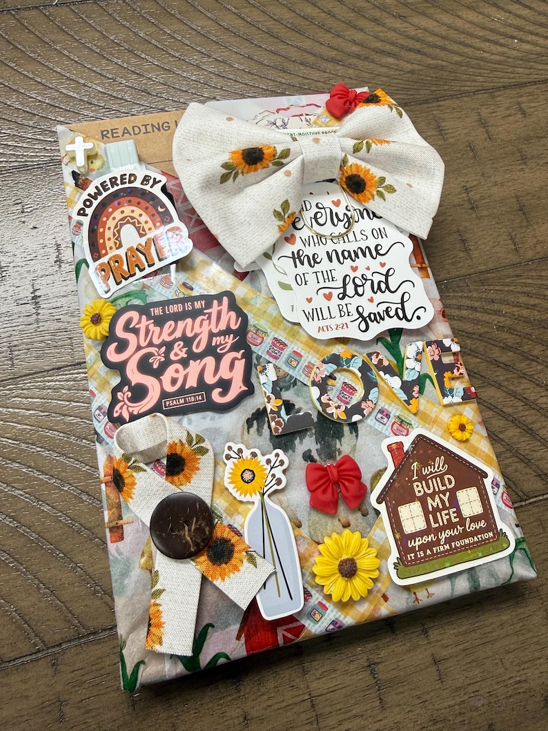 May include: A rectangular devotional book decorated with sunflower-patterned fabric bows, religious stickers, and cutouts. Text includes "Powered by Prayer," "The Lord is my Strength & my Song," "Everyone who calls on the name of the Lord will be saved," and "I will build my life upon your love."