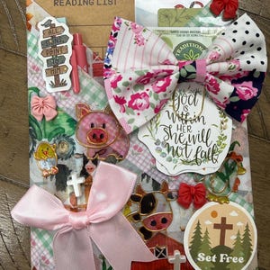 May include: A decorative reading list notebook adorned with a large floral and polka dot fabric bow, a pink satin ribbon, and various stickers including a pig, a cow, a cross, and text reading "God is within her, she will not fall" and "Set Free".