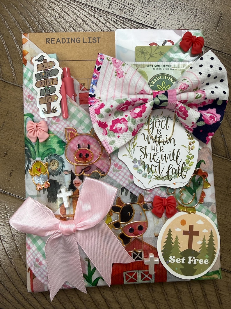 May include: A decorative reading list notebook adorned with a large floral and polka dot fabric bow, a pink satin ribbon, and various stickers including a pig, a cow, a cross, and text reading "God is within her, she will not fall" and "Set Free".