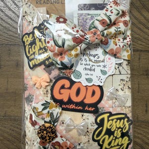 May include: A packaged set featuring a floral fabric bow, stickers with text including "Be the light," "God is within her," and "Jesus is King," a small peach-coloured bow, a black and gold patterned button, and a small white cross charm.