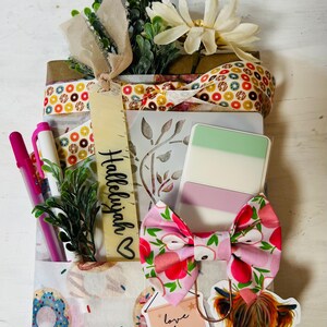 May include: A gift package wrapped in donut-patterned paper, tied with a ribbon featuring colourful circles. It includes a bookmark labelled Hallelujah, a pink apple-print bow, a sticker of a highland cow, and a tag reading love them anyway Luke 23:34.