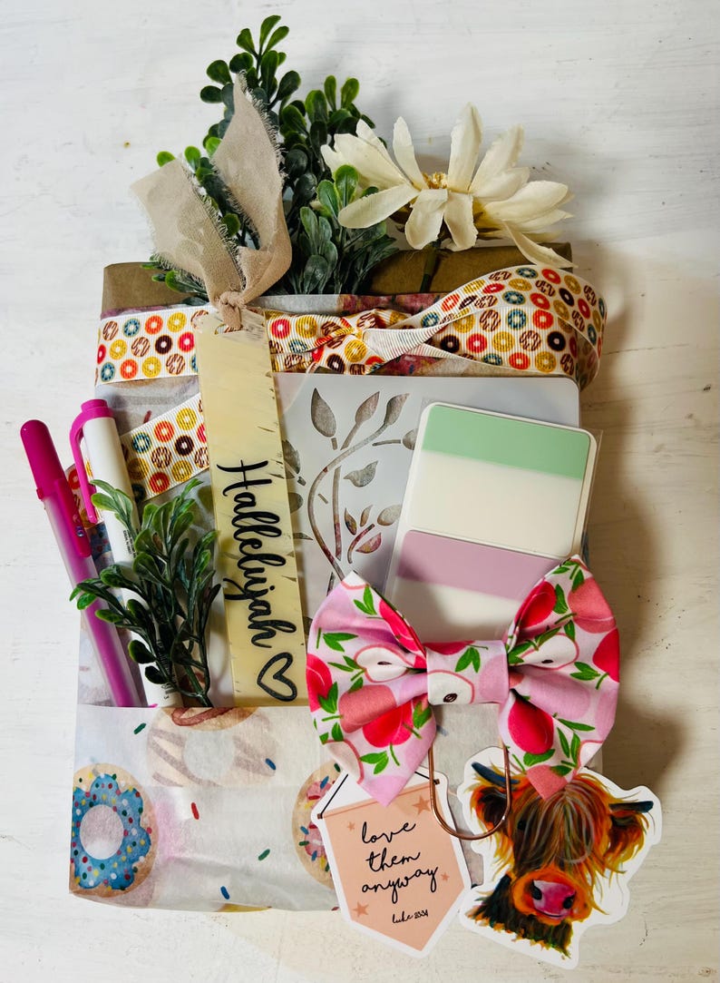 May include: A gift package wrapped in donut-patterned paper, tied with a ribbon featuring colourful circles. It includes a bookmark labelled Hallelujah, a pink apple-print bow, a sticker of a highland cow, and a tag reading love them anyway Luke 23:34.