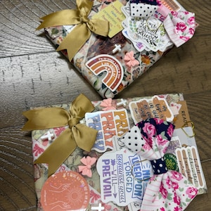 May include: Two rectangular items wrapped in floral paper, each adorned with a gold satin ribbon bow, a fabric bow featuring navy blue and pink floral patterns, small pink bow embellishments, white cross charms, and various religious stickers.