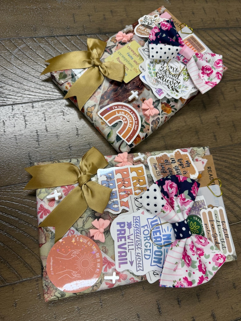 May include: Two rectangular items wrapped in floral paper, each adorned with a gold satin ribbon bow, a fabric bow featuring navy blue and pink floral patterns, small pink bow embellishments, white cross charms, and various religious stickers.