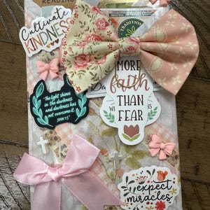 May include: A decorative package featuring a floral and gingham patterned paper base, adorned with a large fabric bow in cream and pink. It includes stickers with text such as Cultivate Kindness, More Faith Than Fear, and Expect Miracles, plus small pink bows and white cross charms.