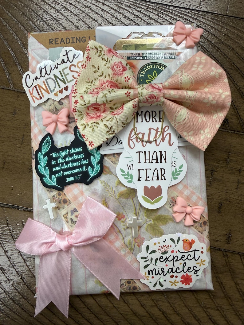 May include: A decorative package featuring a floral and gingham patterned paper base, adorned with a large fabric bow in cream and pink. It includes stickers with text such as Cultivate Kindness, More Faith Than Fear, and Expect Miracles, plus small pink bows and white cross charms.