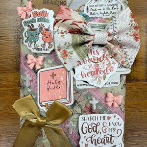 May include: A rectangular package wrapped in floral paper featuring a fabric bow, gold ribbon, and various stickers. Stickers include text such as Build Each Other, Holy Bible, His wounds we are healed, and Search Me God, and My Heart Psalm 139:23.