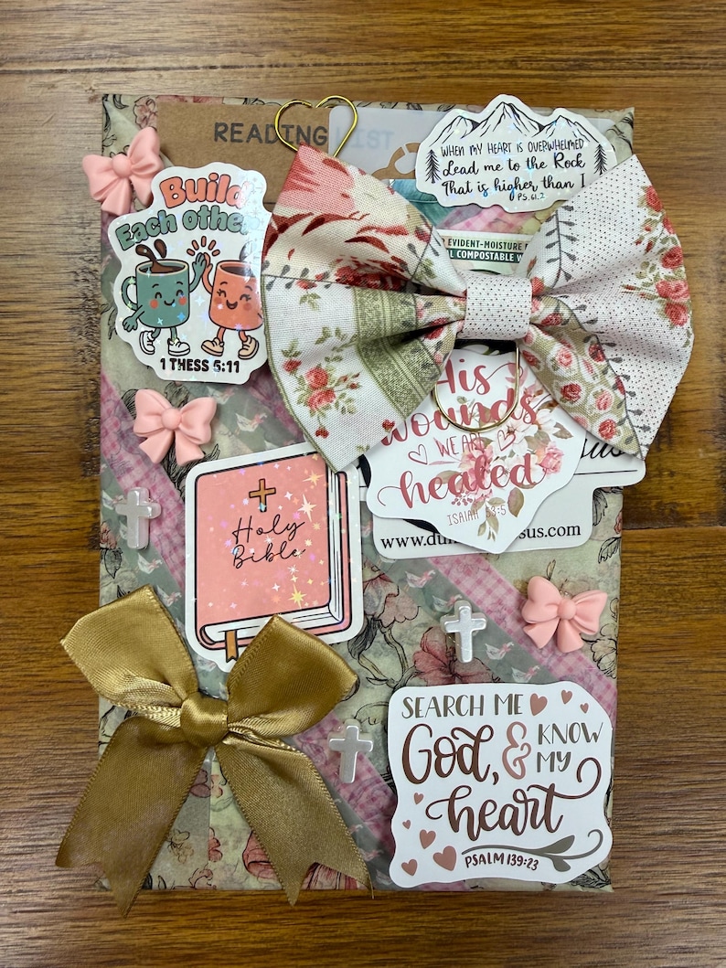 May include: A rectangular package wrapped in floral paper featuring a fabric bow, gold ribbon, and various stickers. Stickers include text such as Build Each Other, Holy Bible, His wounds we are healed, and Search Me God, and My Heart Psalm 139:23.