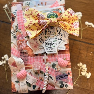May include: A rectangular gift box wrapped in floral paper featuring a yellow fabric bow, a tea bag, and various stickers with religious text including "forgive one another" and "worship". It is decorated with pink pumpkin charms, a cross bead strand, and ribbon.