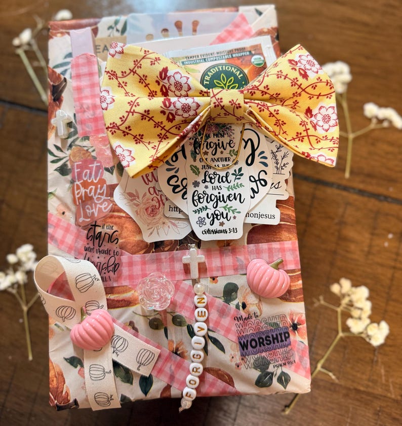 May include: A rectangular gift box wrapped in floral paper featuring a yellow fabric bow, a tea bag, and various stickers with religious text including "forgive one another" and "worship". It is decorated with pink pumpkin charms, a cross bead strand, and ribbon.