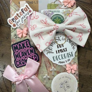 May include: A rectangular package wrapped in floral paper featuring a large cream bow with pink ribbon patterns, a pink satin bow, and several stickers. Text includes Grateful for Jesus, Make Heaven Crowded, He must increase but I must decrease John 3:30, and It is well with my soul. Small cross charms and bow-shaped embellishments are attached.