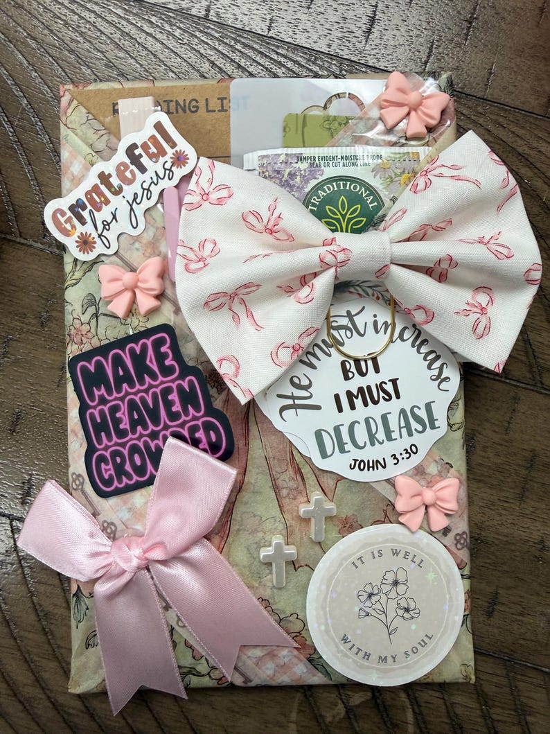 May include: A rectangular package wrapped in floral paper featuring a large cream bow with pink ribbon patterns, a pink satin bow, and several stickers. Text includes Grateful for Jesus, Make Heaven Crowded, He must increase but I must decrease John 3:30, and It is well with my soul. Small cross charms and bow-shaped embellishments are attached.