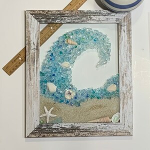 May include: A framed artwork featuring a blue glass wave with white seashells and a starfish on a sandy beach background.