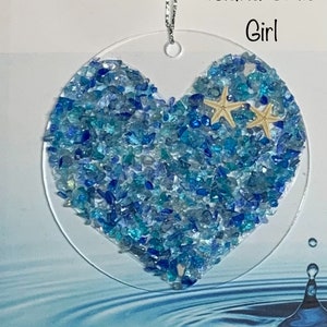 May include: A heart-shaped ornament made of blue and green glass pieces. The ornament is surrounded by a clear circle and has two small starfish on top. The ornament is sitting on a blue water background with ripples.