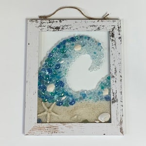 May include: A whitewashed wood framed wall art piece featuring a blue and white wave made of sea glass and seashells. The wave is set against a white background with a sandy beach at the bottom.