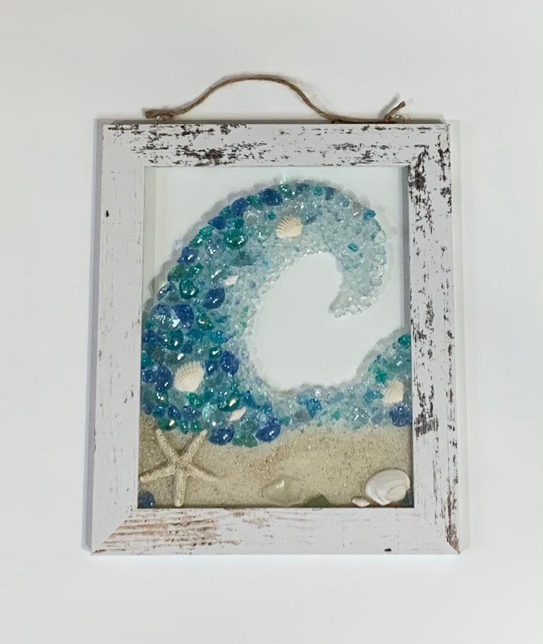 May include: A whitewashed wood framed wall art piece featuring a blue and white wave made of sea glass and seashells. The wave is set against a white background with a sandy beach at the bottom.