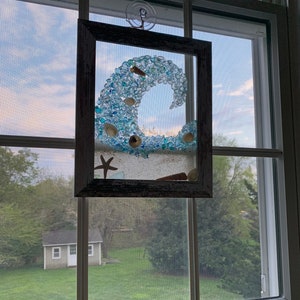 May include: A framed piece of art featuring a blue wave made of glass beads, seashells, and a starfish. The wave is set against a white sandy background. The frame is made of wood and has a distressed white finish.