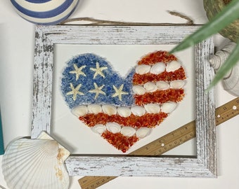 Op de afbeelding: Een wit houten frame met een hartvormige Amerikaanse vlag gemaakt van schelpen en glazen kralen. De vlag is rood, wit en blauw. Het frame is verweerd en heeft een rustieke uitstraling.