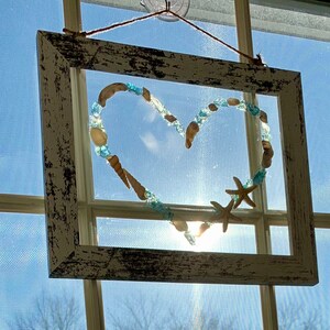 K&ouml;nnte beinhalten: Ein wei&szlig;er Holzrahmen mit einem herzf&ouml;rmigen Design aus Muscheln und blauen Glasperlen. Das Herz ist von einem wei&szlig;en Hintergrund umgeben und der Rahmen h&auml;ngt an einem Fenster.