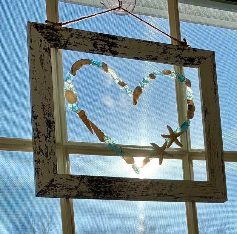 K&ouml;nnte beinhalten: Ein wei&szlig;er Holzrahmen mit einem herzf&ouml;rmigen Design aus Muscheln und blauen Glasperlen. Das Herz ist von einem wei&szlig;en Hintergrund umgeben und der Rahmen h&auml;ngt an einem Fenster.