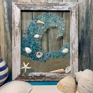 May include: A framed seashell and glass mosaic artwork depicting a wave crashing on a sandy beach. The frame is whitewashed wood with a distressed finish. The wave is made of blue and green glass pieces, and the sand is represented by a layer of glitter.