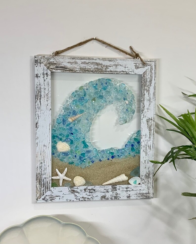 May include: A framed wall art piece featuring a blue wave made of sea glass and seashells. The frame is whitewashed wood with a distressed finish.