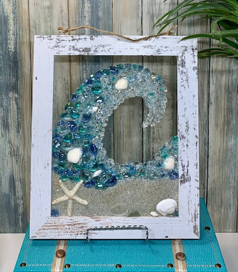 May include: A white framed wall art piece featuring a blue and green glass mosaic wave with white seashells and a starfish on a white sand base.