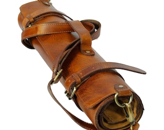May include: Rolled brown leather knife roll with multiple straps and brass-colored hardware. The roll is designed to hold knives and other culinary tools. The leather has a rich, textured appearance, and the straps secure the roll.