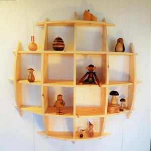 May include: A light-coloured wooden shelf unit with a circular design, featuring multiple cubby spaces. The shelves display various wooden figurines, including a monkey, a penguin, and a mushroom. A candle and a decorative egg are also visible.