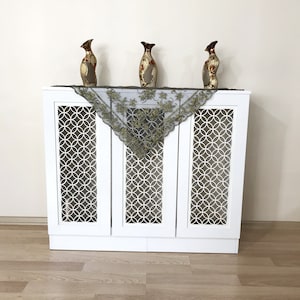 May include: A white cabinet with a geometric circle design on the doors. Three decorative vases and a dark green lace scarf adorn the top. The vases are gold, brown, and red, adding a touch of elegance.