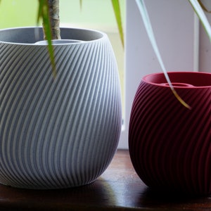 Puede incluir: Dos macetas decorativas con un diseño de ranuras en espiral. Una es de color gris claro y la otra de color rojo intenso. Las macetas están sobre una superficie de madera cerca de una ventana, con follaje verde visible.