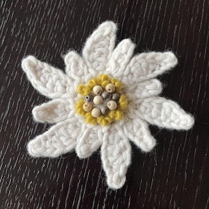 Könnte beinhalten: Eine gehäkelte Edelweißblüte mit cremeweißen Garnblütenblättern in Sternform. Die Mitte besteht aus strukturierten, senfgelben Garnschlingen, die mit kleinen, runden, hellbeigen und braunen Holzperlen verziert sind.